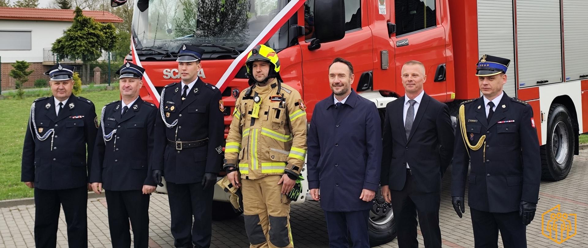 Zdjęcie przedstawia uczestników uroczystości przekazania ciężkiego samochodu ratowniczo-gaśniczego GCBA 5/32 Scania P380. Przekazujący oficjele wraz z przedstawicielami jednostki OSP stoją przed przekazanym samochodem gaśniczym.