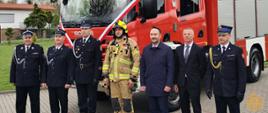 Zdjęcie przedstawia uczestników uroczystości przekazania ciężkiego samochodu ratowniczo-gaśniczego GCBA 5/32 Scania P380. Przekazujący oficjele wraz z przedstawicielami jednostki OSP stoją przed przekazanym samochodem gaśniczym.