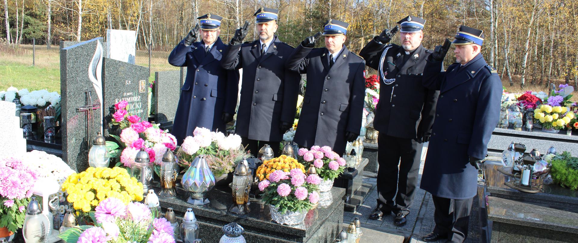Zdjęcie przedstawia funkcjonariuszy Państwowej Straży Pożarnej oraz kapelana ubranych w mundury wyjściowe, którzy oddają honor nad grobem zmarłego strażaka. W tle widoczne są nagrobki na których znajdują się znicze oraz kwiaty.