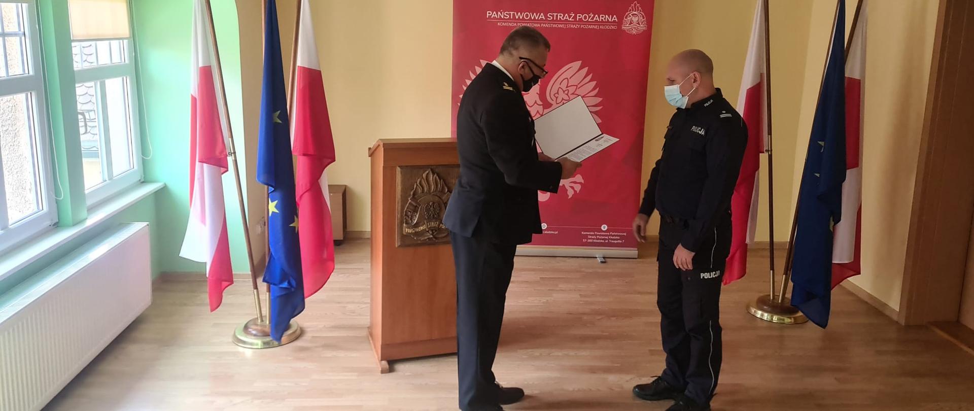 Komendant Powiatowy Państwowej Straży Pożarnej w Kłodzku wręcza funkcjonariuszowi policji list pochwalny na Sali Konferencyjnej. 