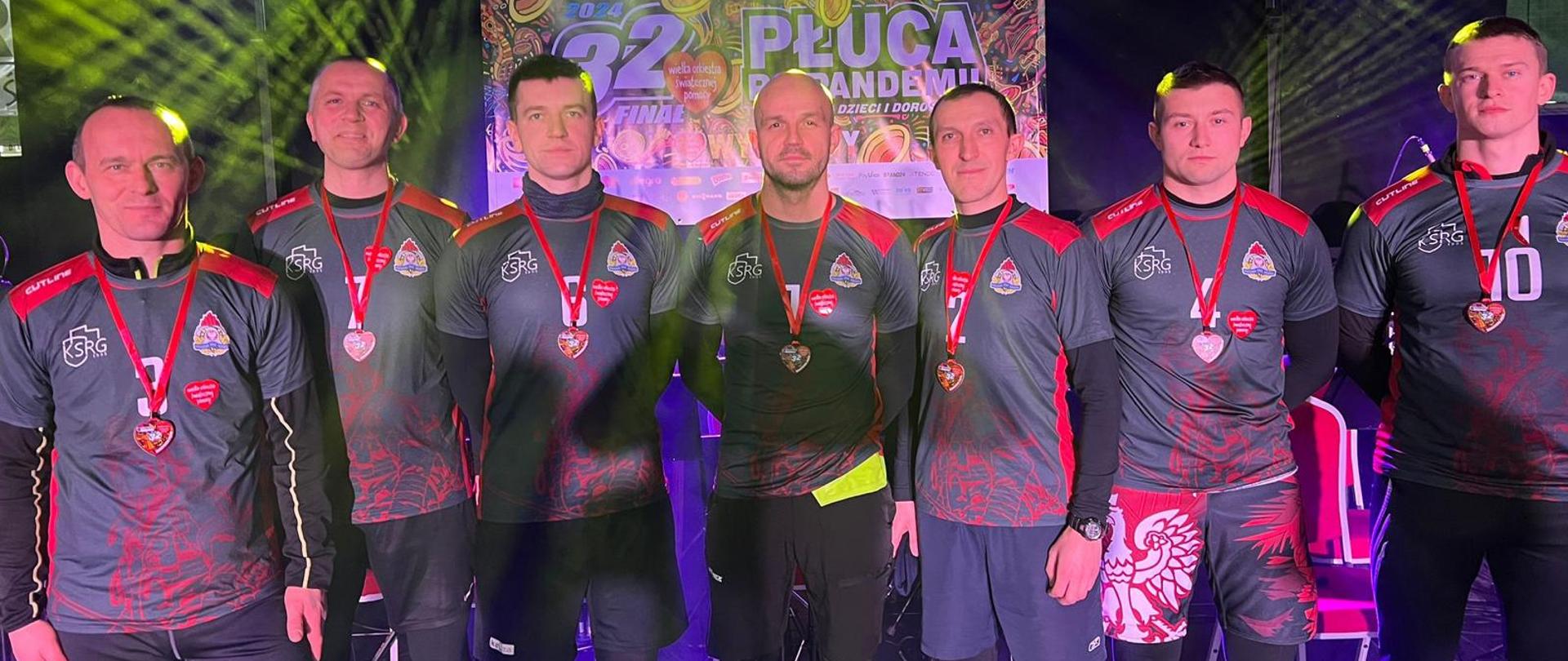 Na zdjęciu siedmiu strażaków z Komendy Powiatowej Państwowej Straży Pożarnej w Gorlicach w strojach sportowych z medalami na szyi na scenie 32 Finału Wielkiej Orkiestry Świątecznej Pomocy na hali Ośrodka Sportu i Rekreacji w Gorlicach po przebiegnięciu biegu „Orkiestrowa Piątka” zbierając pieniądze na Wielką Orkiestrę Świątecznej Pomocy. 