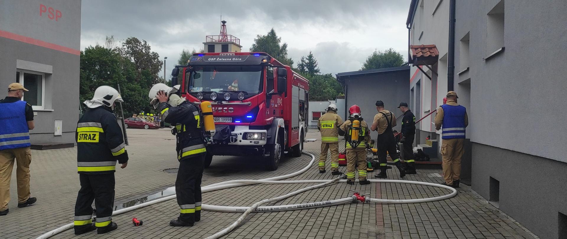 Zdjęcie przedstawia ćwiczenia strażackie. Na pierwszym planie znajduje się czerwony wóz strażacki OSP Żelazna Góra, do którego podłączone są węże gaśnicze. Wokół pojazdu widocznych jest wielu strażaków – część z nich ubrana jest w pełne umundurowanie bojowe z aparatami powietrznymi, a inni są w kamizelkach organizacyjnych.
Strażacy przygotowują się do działania – jeden zakłada hełm, inny sprawdza sprzęt. Po lewej stronie w tle widać drugi pojazd oraz manekina lub atrapę samochodu, co sugeruje, że ćwiczenia mogą obejmować symulację wypadku lub pożaru. Pogoda jest pochmurna, a teren wybrukowany i przylega do budynków jednostki. Zdjęcie ukazuje profesjonalne przygotowanie do akcji ratowniczo-gaśniczej.
