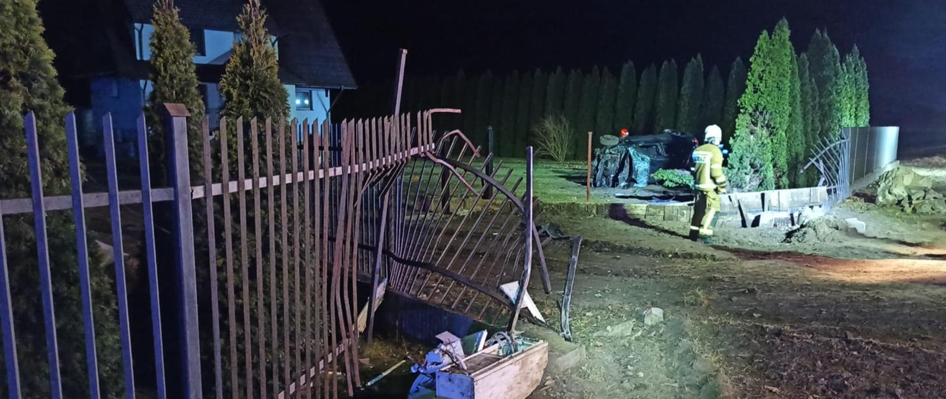 Na posesji znajduje się uszkodzony samochód wraz z ogrodzeniem.