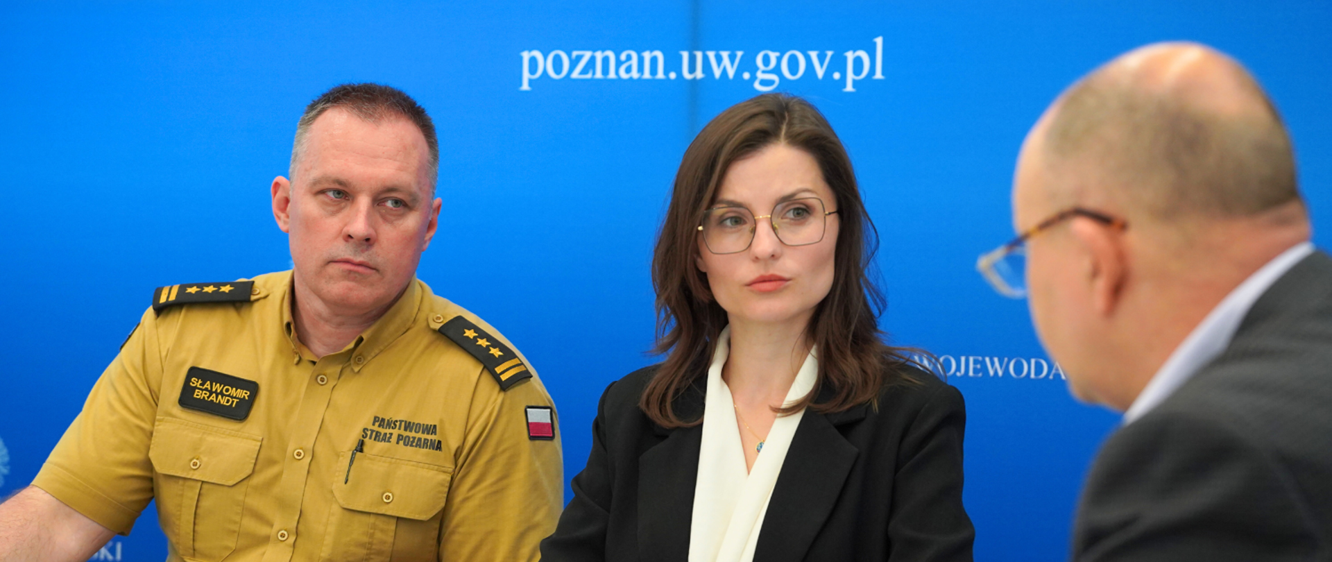 Zastępca komendanta wojewódzkiego PSP, wojewoda i wicewojewoda oraz urzędnicy zajęli miejsca przy stole i słuchają wideokonferencyjnego wystąpienia