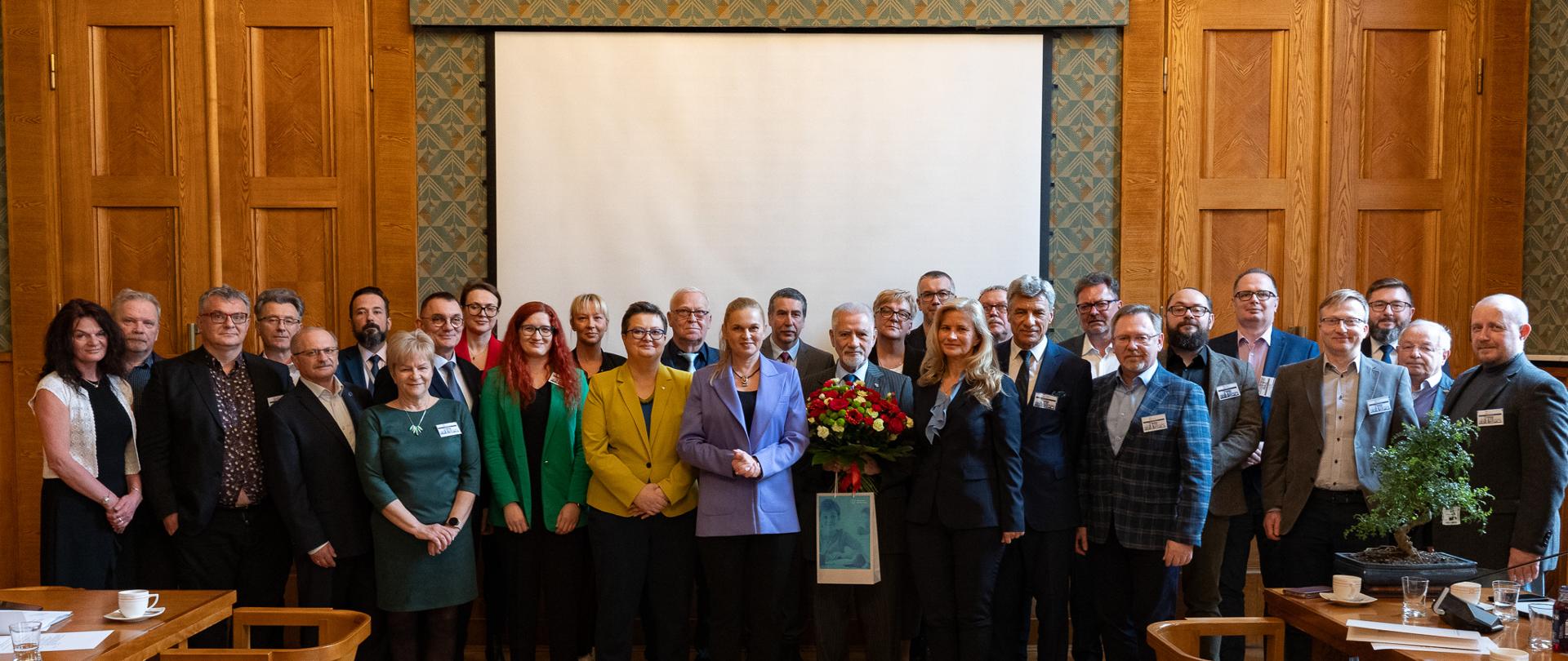 Zdjęcie zbiorowe, na sali pod ścianą stoi duża grupa ludzi.