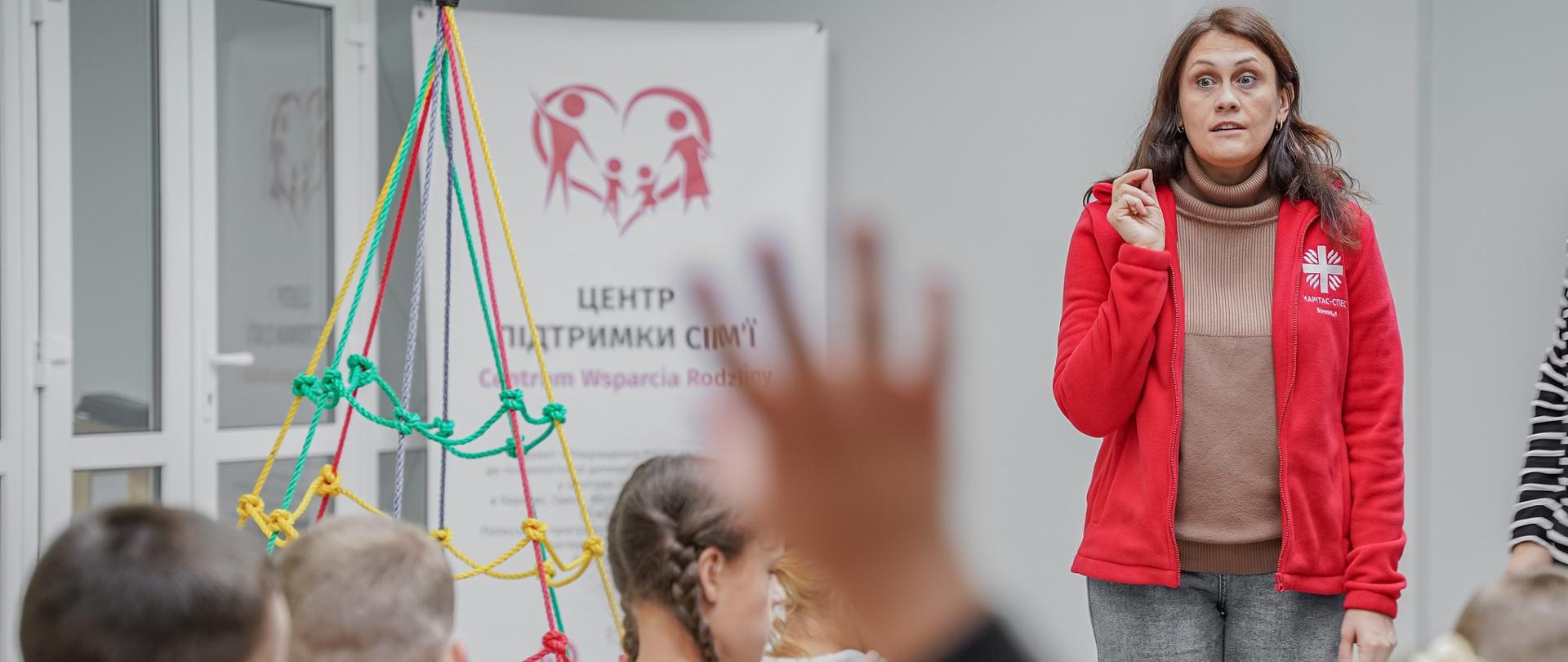 Kobieta w czerwonym polarze z napisem Caritas prowadzi zajęcia dla grupki dzieci odwróconych tyłem na zdjęciu, widać 1 uniesioną rękę