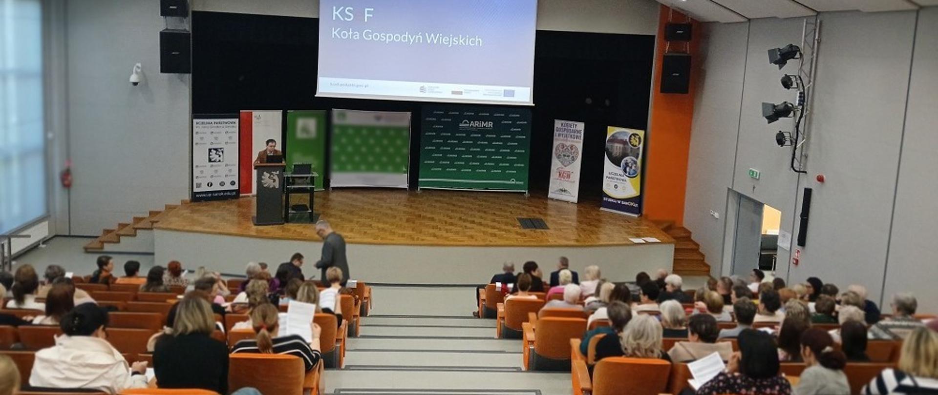 W sali siedzą licznie zgromadzeni uczestnicy szkolenia. Na ekranie wyświetlana jest prezentacja.