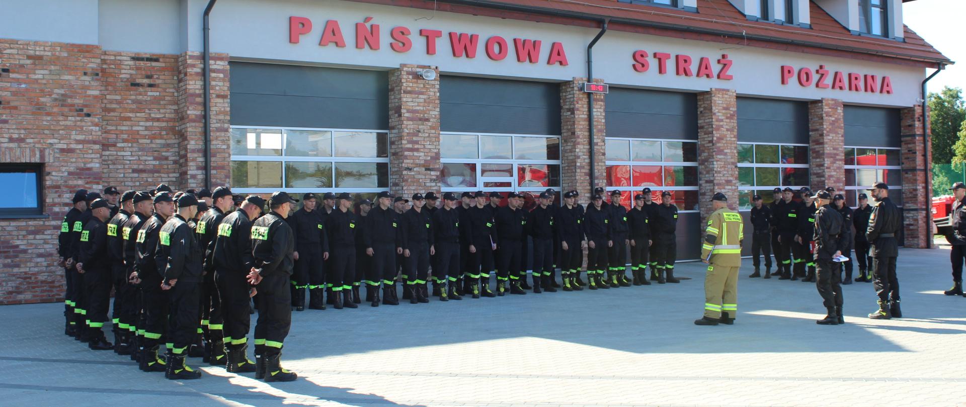 Przed budynkiem Komendy Powiatowej Państwowej Straży Pożarnej Dowódca Kompanii przeprowadza odprawę ze strażakami wchodzącymi w skład kompanii, stojącymi w dwuszeregu.