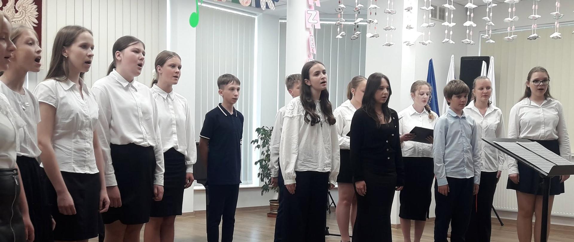 Grupa dziewcząt i chłopców stoi w sali koncertowej. Przed nimi ustawiony pulpit z nutami. W tle okna zasłonięte żaluzjami.