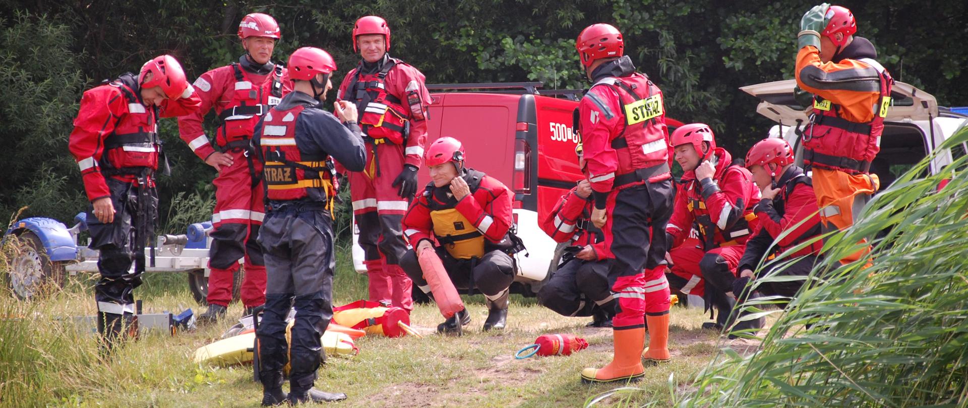 Zdjęcie przedstawia uczestników szkolenia w trakcie instruktażu odnośnie sposobów
prowadzenia działań na akwenach wodnych. Strażacy stoją na brzegu. Ćwiczący ubrani są w suche skafandry ratownicze oraz kamizelki asekuracyjne. Głowy zabezpieczone maja kaskami ochronnymi z przeznaczeniem do ratownictwa wodnego.
