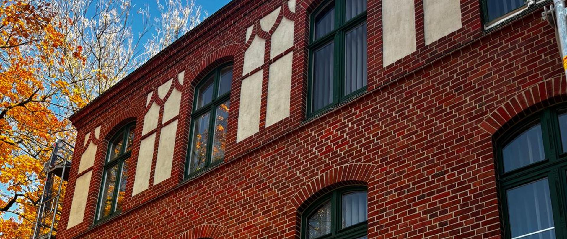 Fragment ceglanej ściany Państwowej Szkoły Muzycznej w Szczecinku po remoncie. Odnowiona, czysta cegła w kolorze czerwonym oraz 8 okien z zielonymi framugami i 3 okna z białymi framugami. W tle niebo i fragment drzewa z pomarańczowymi liśćmi.