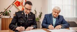 Funkcjonariusz PSP w czarnym mundurze galowym oraz mężczyzna w niebieskim garniturze, którzy wspólnie podpisują dokumenty przy stole w gabinecie; w tle widoczny sztandar jednostki z napisem „Bogu na chwałę, ludziom na pożytek” oraz symbole strażackie. Mężczyźni mają głowy pochylone nad podpisywanymi dokumentami.