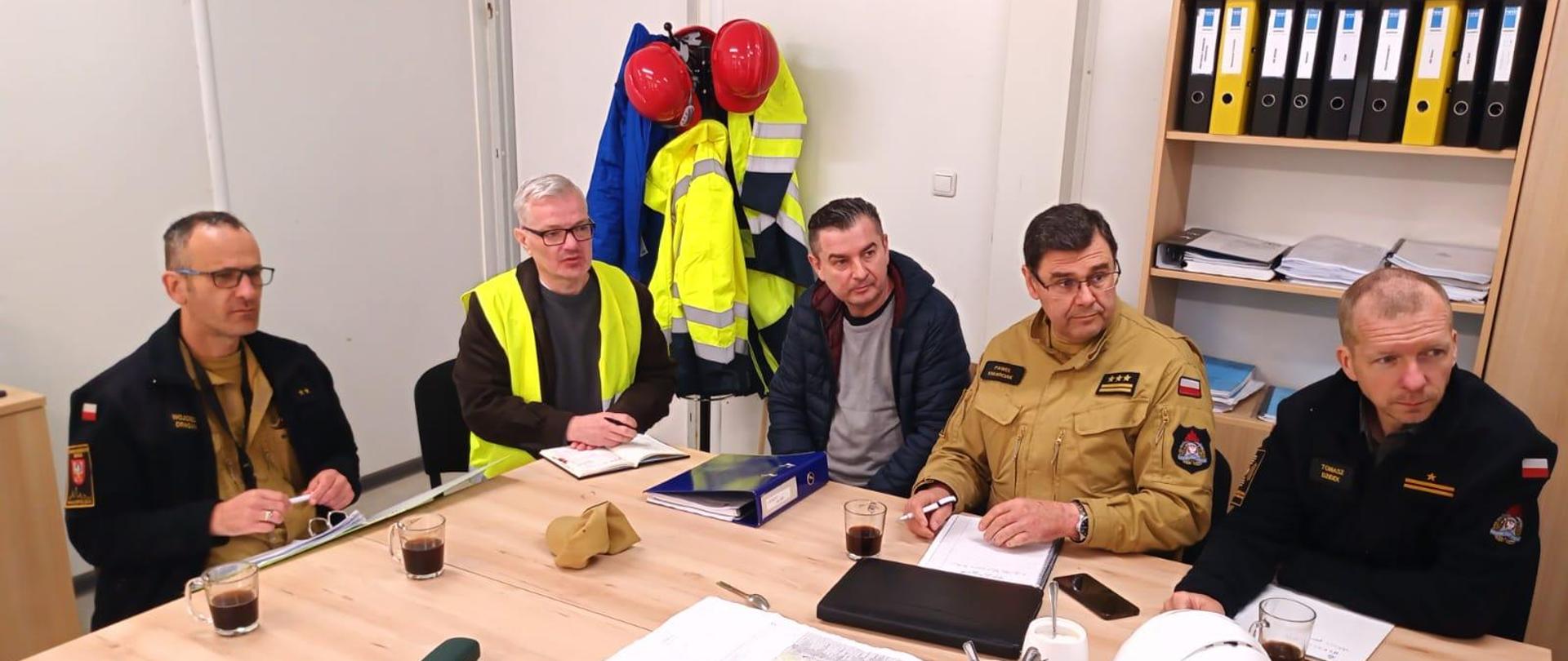 Strażacy oraz pracownicy budowy siedzą w biurze przy stole na którym znajduje się dokumentacja projektowa.