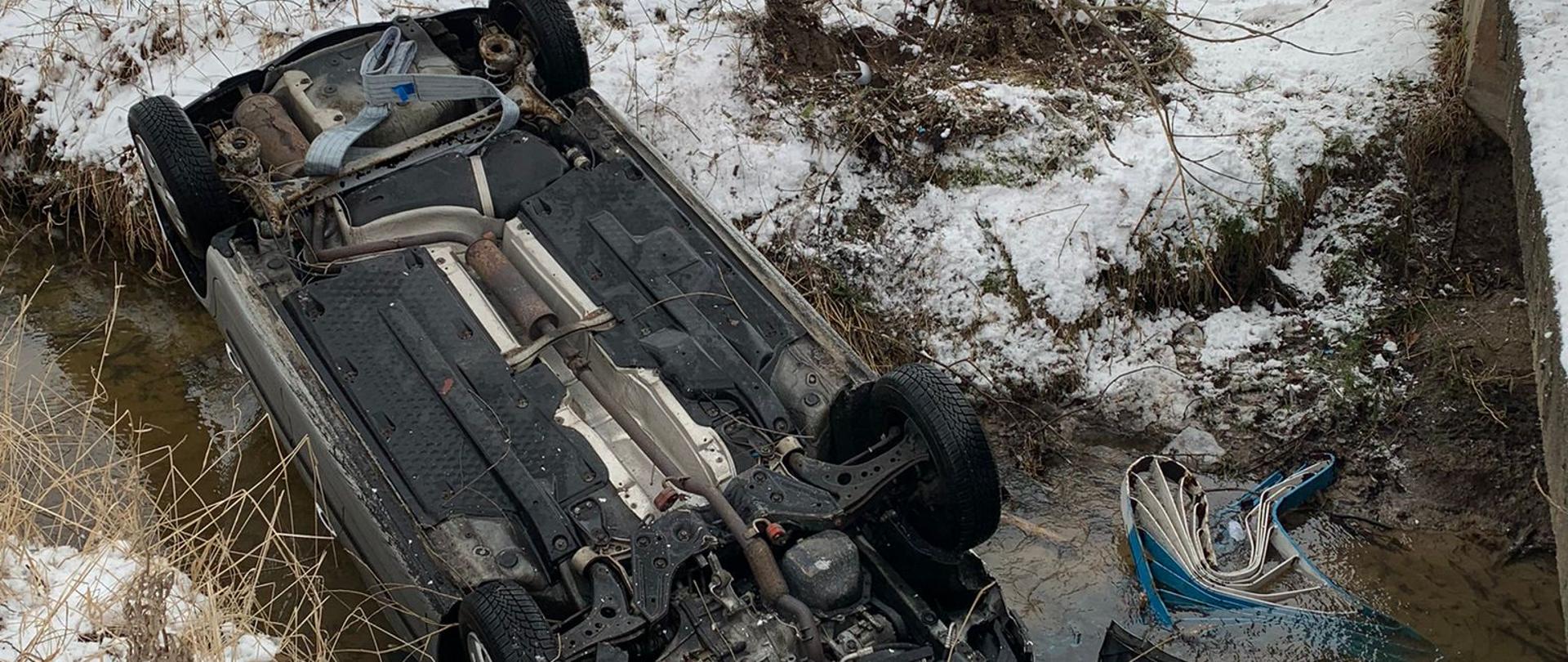 Samochód osobowy leży na dachu w rzece. Z tył trzech strażaków na ziemi leży śnieg.