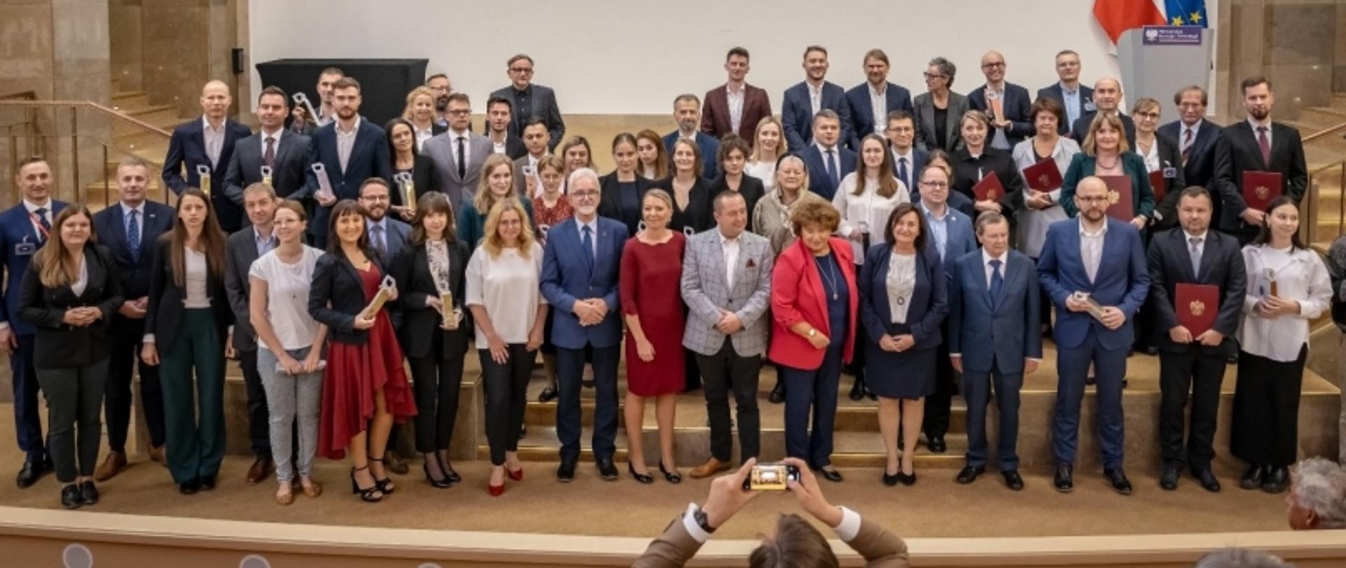 Grupowe zdjęcie uczestników ceremonii z certyfikatami, ustawionych na schodach, w tle flagi.