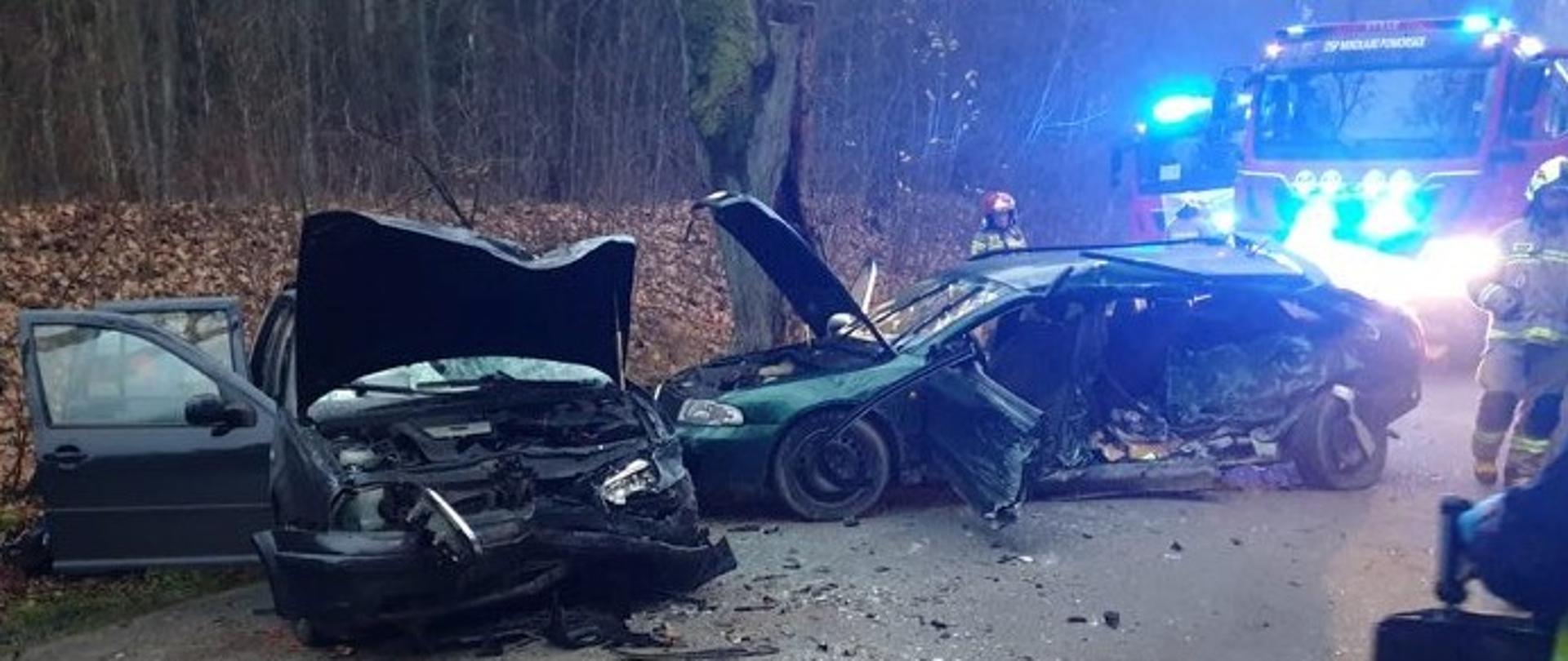 kadr przedstawia drogę w lesie, na środku pasa dwa rozbite auta, za wrakami widać strażaka i wóz straży pożarnej
