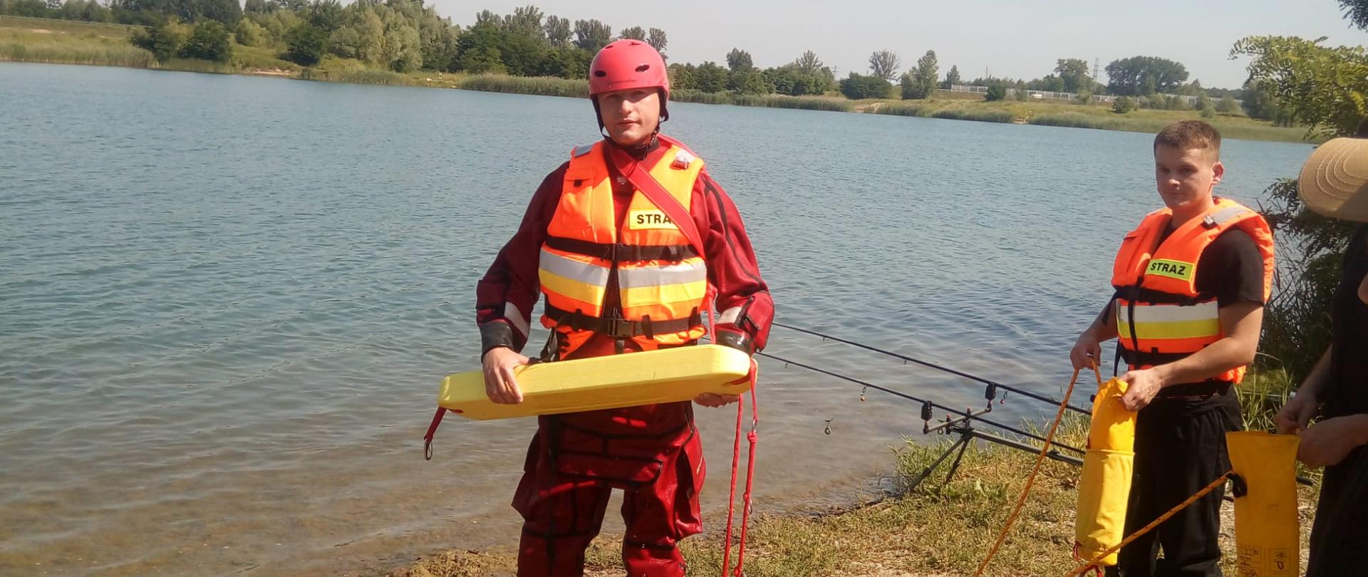 Zdjęcie przedstawia ćwiczenia z zakresu ratownictwa wodnego. Na zdjęciu widać strażaka ubranego w czerwony kombinezon do pracy w wodzie. Na głowie ma czerwony kask. W ręku trzyma pas typu węgorz.. Obok strażak znajduje się drugi strażak ubrany w kamizelkę ratunkową i trzyma w ręku rzutkę ratowniczą