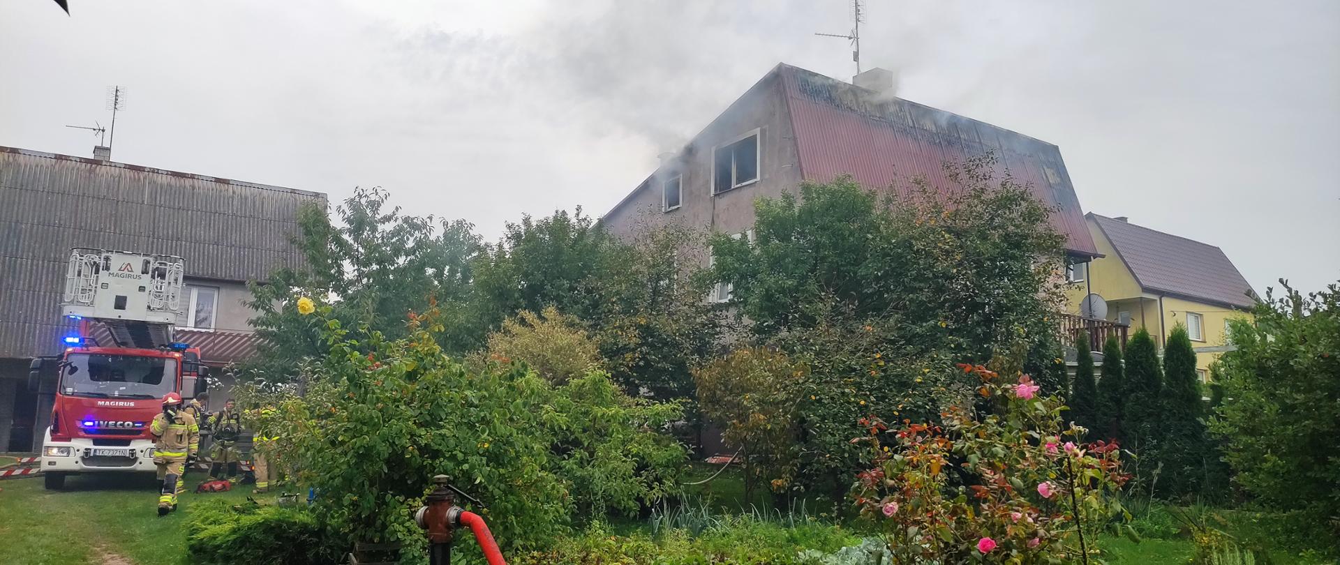 Zdjęcie przedstawia budynek, w którym wybuchł pożar. Dym unosi się ze zniszczonego dachu. Po lewej stronie zaparkowany stoi samochód pożarniczy.
