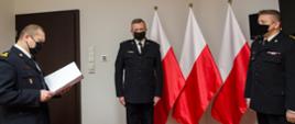 Zdjęcie przedstawia trzech strażaków w umundurowaniu wyjściowym stojących na tle trzech flag Polski. Po lewej stronie strażak czyta akt mianowania. Strażak w środku stoi na baczność. Z prawej strony stoi mazowiecki komendant wojewódzki PSP.