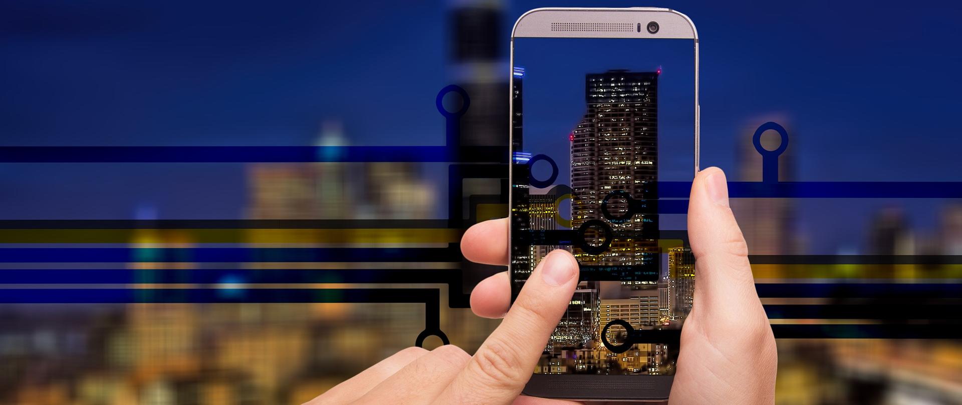 Dłonie trzymające smartfona. W tle miejskie wieżowce.