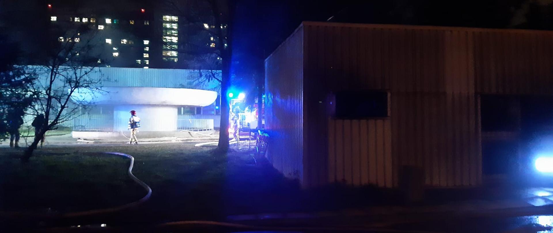 Zdjęcie przedstawia dwa budynki, oraz pojazd ratowniczy z włączonym niebieskim oświetleniem błyskowym, oraz kierującego działaniem ratowniczym PSP.