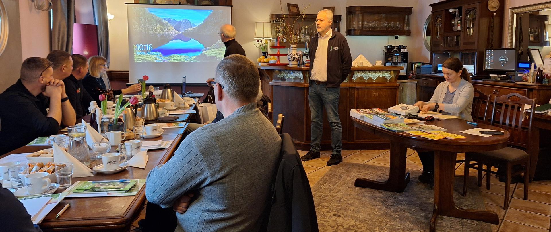 Uczestnicy siedzibą z obu stron stołu z lewej strony, po prawej przy stole naprzeciw siedzi jedna osoba. W głębi zdjęcia ekran z prezentacją, pośrodku stoi inspektor 