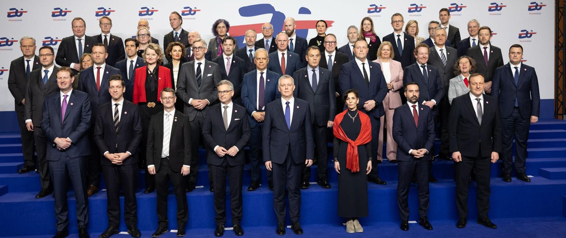 Ministrowie pozują do family photo na granatowych schodach. Tło stanowi ścianka z logotypem polskiej prezydencji w Radzie UE.