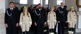 Strażacy OSP w mundurach wyjściowych stoją na przemian ze strażakami z młodzieżowej drużyny pożarniczej w ubraniach koszarowych koloru beżowego.