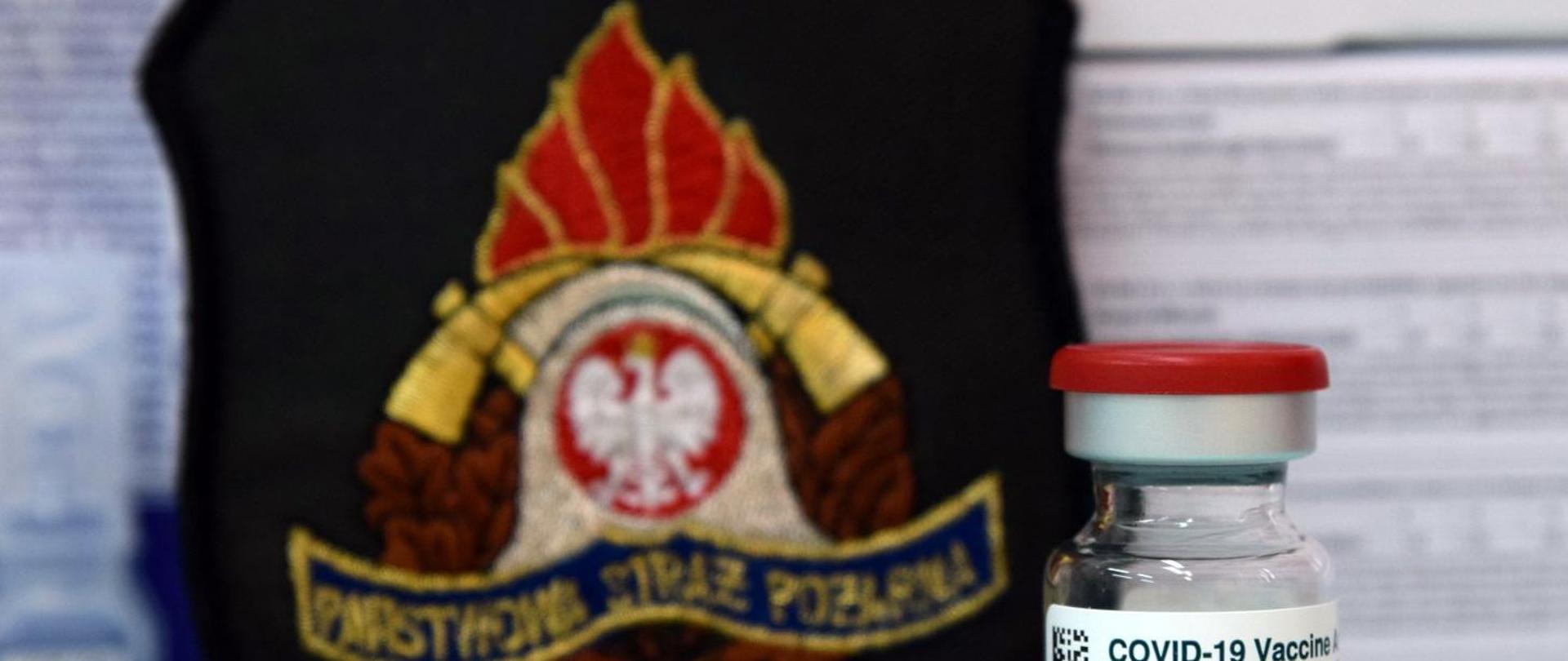 Na zdjęciu emblemat z logo Państwowej Straży Pożarnej a przed nim fiolka medyczna z napisem COVID-19 Vaccine