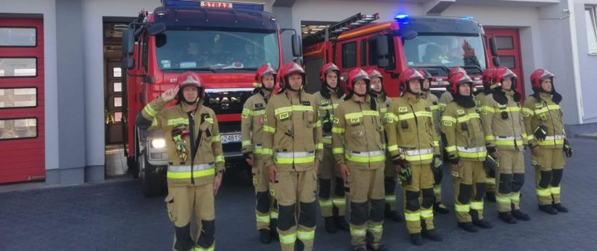 Zdjęcie wykonane przed budynkiem Komendy. Centralny punkt stanowią stojący przed Komendą w dwuszeregu strażacy. Strażacy mają piaskowe munduru i czerwone hełmy. W tle budynek Komendy i pojazdy gaśnicze.