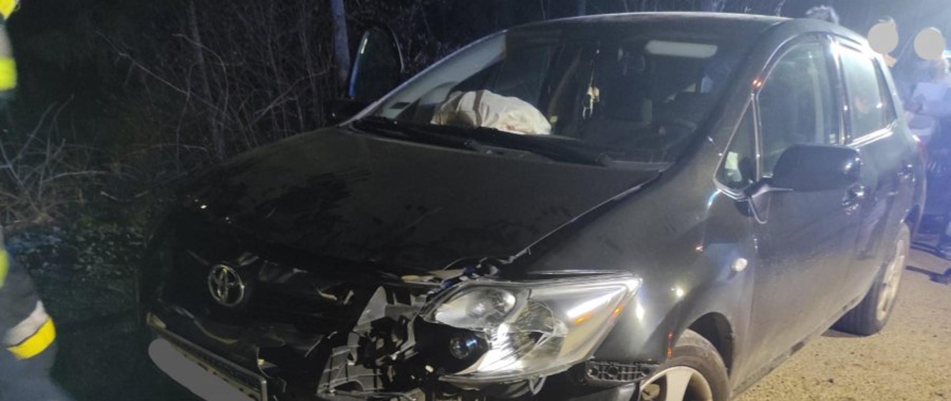 Zdjęcie przedstawia rozbity czarny samochód osobowy. W dalszym planie widzimy osoby postronne obserwujące zdarzenie. Zdarzenie w porze nocnej