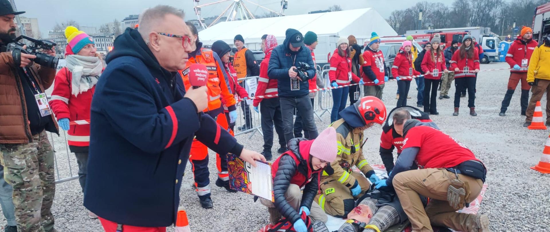 Pokaz ratownictwa w trakcie Finału w Warszawie. Prowadzący wskazuje na ćwiczących ratowników