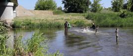 w korycie rzeki stoi dwóch strażaków ubranych w skafandry, zabezpieczają zawodników wchodzących do wody, jeden się przewraca i zanurza się w wodzie po szyję