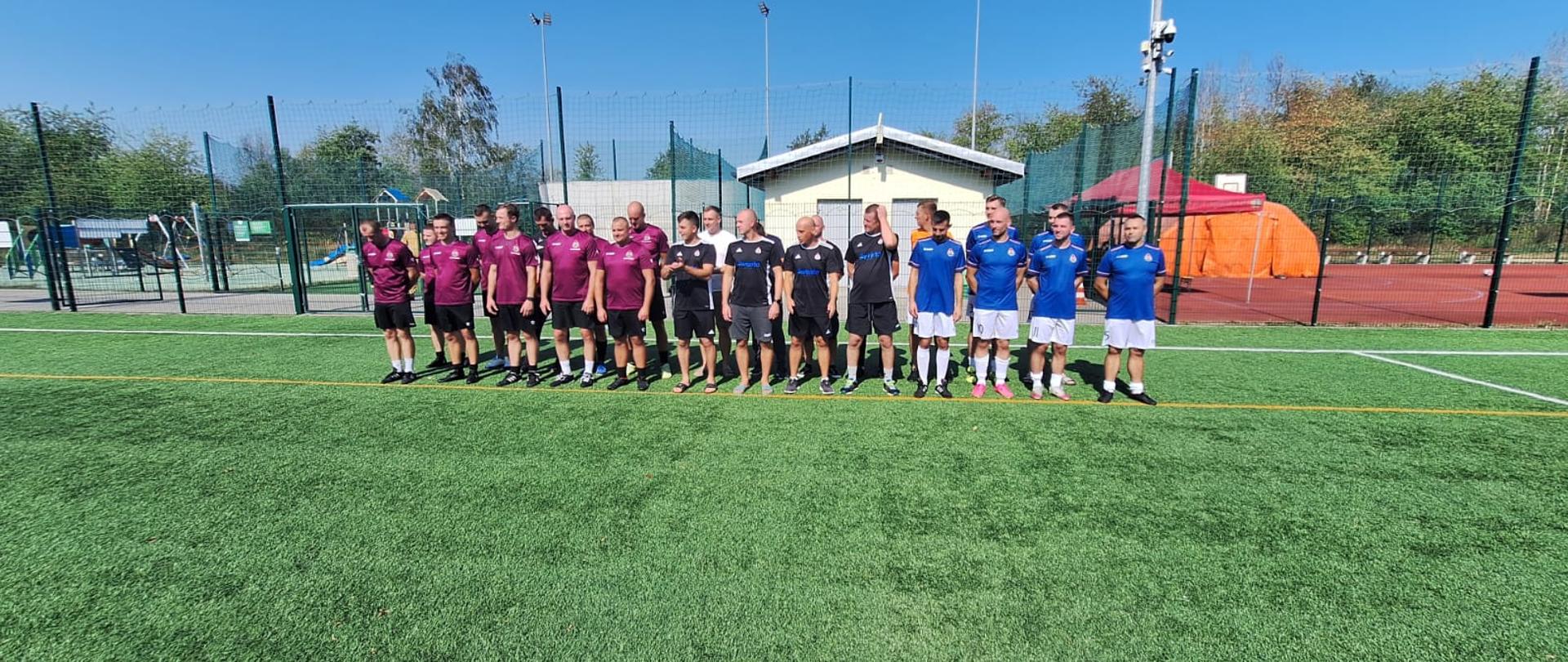 Na zdjęciu widzimy trzy drużyny piłkarskie ustawione w rzędzie na murawie boiska, najprawdopodobniej przed rozpoczęciem turnieju.
Zawodnicy pozują do wspólnego zdjęcia grupowego, co sugeruje oficjalny charakter wydarzenia sportowego.
Szczegóły zdjęcia:
Drużyny:
Po lewej stronie zawodnicy w bordowych koszulkach, czarnych spodenkach – tworzą największą grupę.
W środku znajdują się zawodnicy w czarnych koszulkach z niebieskim napisem.
Po prawej stronie stoi drużyna w niebieskich koszulkach i białych spodenkach.
Tło: Widać ogrodzony teren boiska, plac zabaw, mały budynek (prawdopodobnie zaplecze techniczne lub szatnie) oraz czerwony namiot techniczny i pomarańczowy namiot (mogący pełnić funkcję zaplecza organizacyjnego lub punktu medycznego).