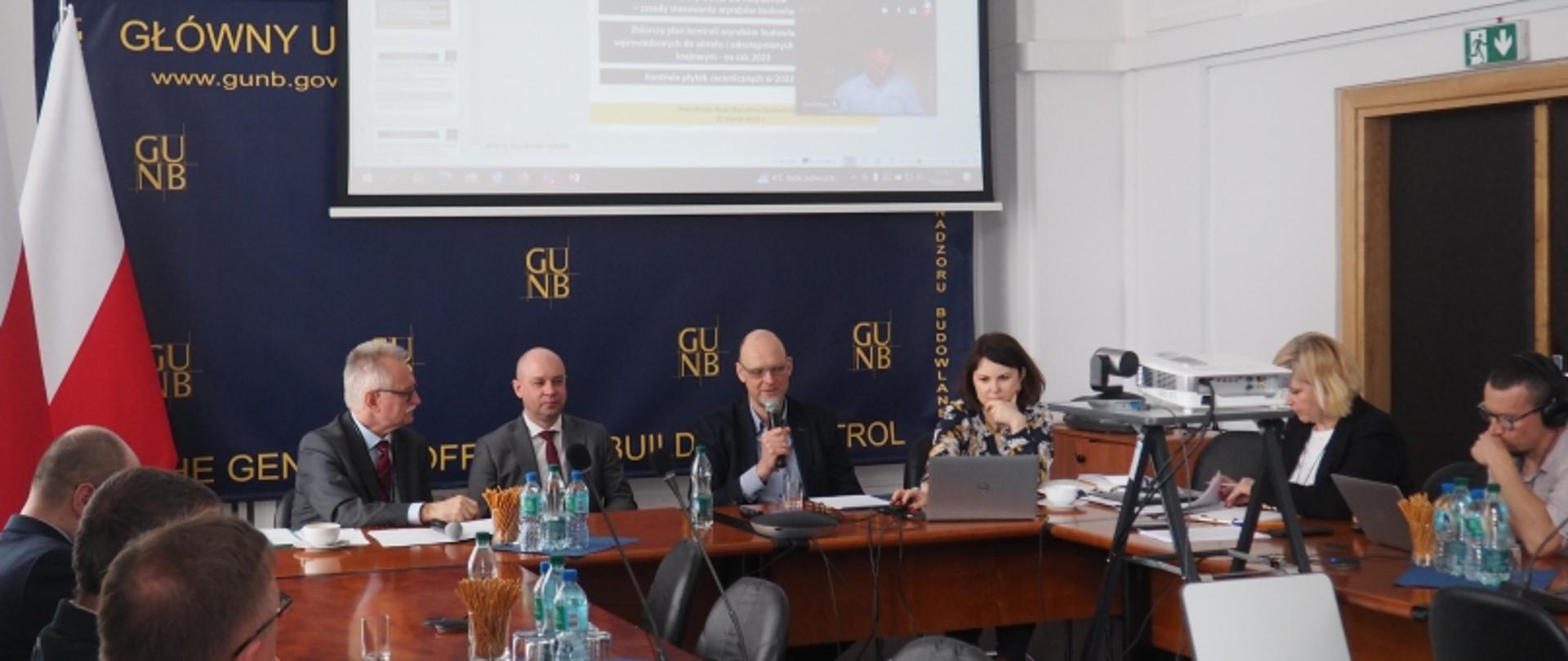 Sala konferencyjna z uczestnikami przy stole, ekranem prezentacyjnym, banerem GUNB i flagą Polski.