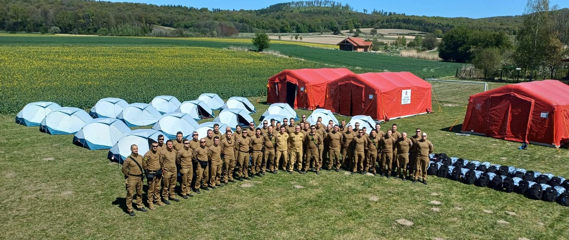 o zdjęcie przedstawia dużą grupę umundurowanych strażaków, pozujących do wspólnej fotografii na otwartym terenie.