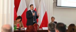 Minister Czarnek stoi za stołem i mówi do mikrofonu, za nim polskie flagi.