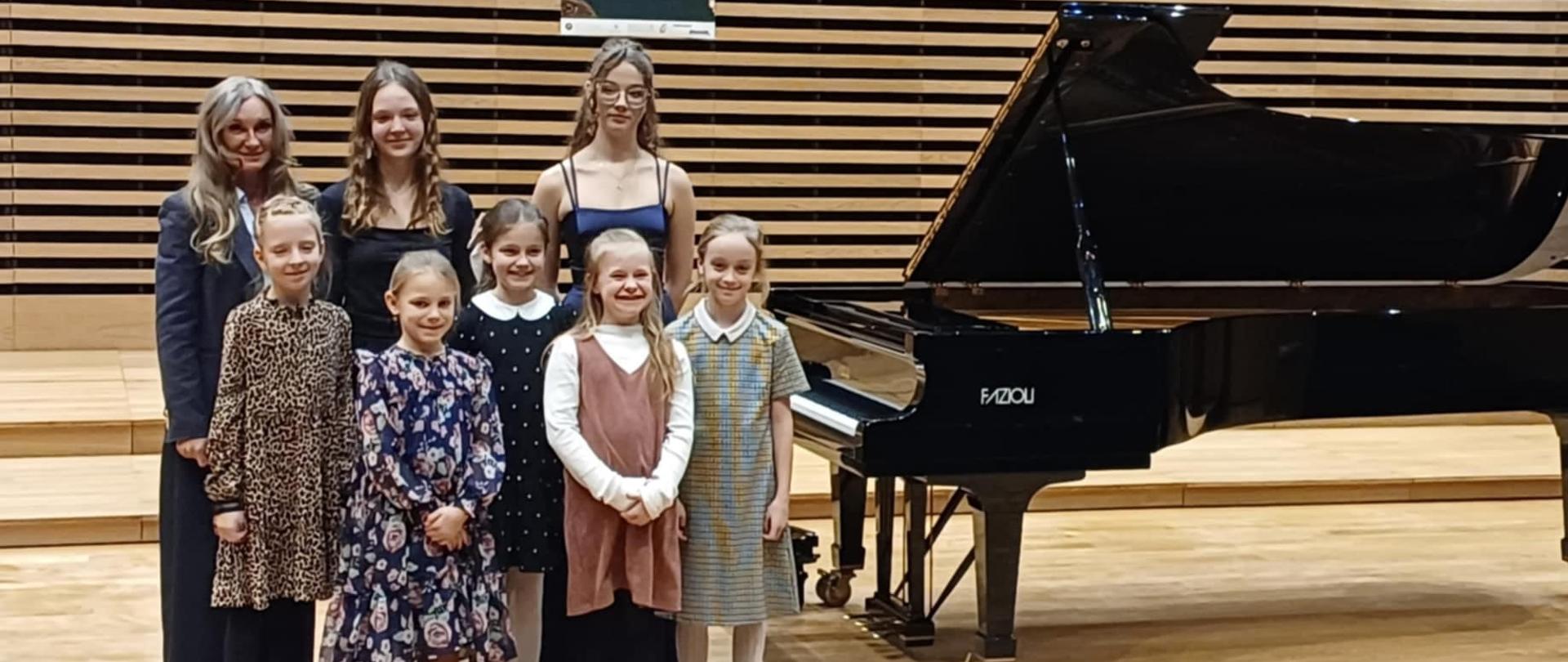 Wszystkie uczennice razem z nauczycielką stoją obok fortepianu na estradzie.