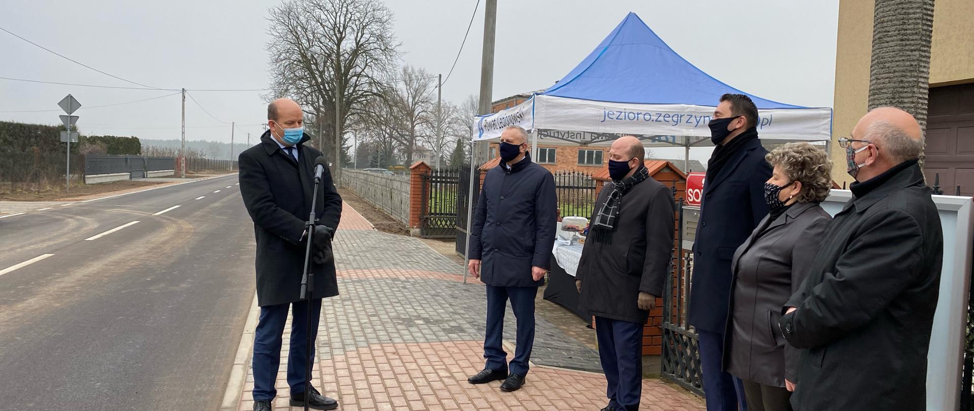 Wojewoda Mazowiecki na drodze, przy mikrofonie, w eleganckim płaszczu, przemawia do piątki osób.