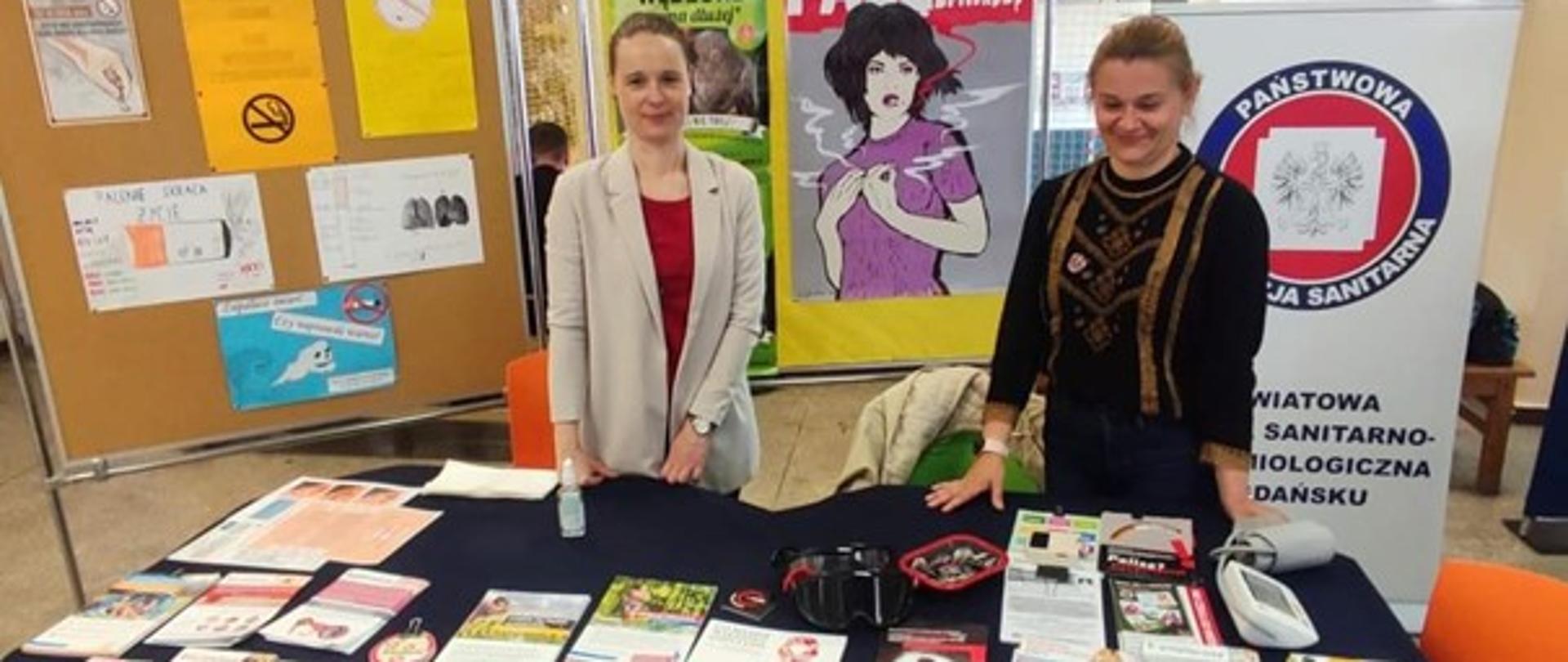 Dwie kobiety stoją przy stoisku z ulotkami informacyjnymi. Za kobietami logo Powiatowej Stacji Sanitarno-Epidemiologicznej w Gdańsku oraz plakaty antynikotynowe.