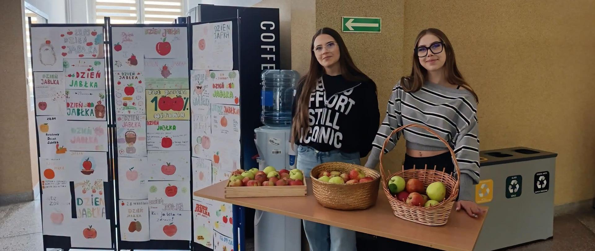 Na zdjęciu w szkolnym korytarzu znajdują się dwie uczennice stojące przy stoisku z jabłkami. Na stole ustawiono kosze i skrzynki wypełnione jabłkami oraz dystrybutor wody. Obok znajduje się ekspozycja z kolorowymi, własnoręcznie wykonanymi plakatami poświęconymi Dniu Jabłka. W tle widoczne są szkolne wnętrza, znak ewakuacyjny oraz pojemniki do segregacji odpadów.
