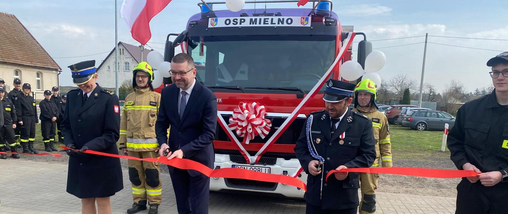 Na zdjęciu widać pododdział strażacki biorący udział w uroczystej zbiórce. W tle stoją samochody pożarnicze.