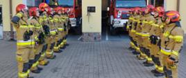 Strażacy stoją na bacznóść przed budynkiem komendy i oddają honor dla poległych strażaków z Ukariny. Za nimi widac 2 pojazdy gaśnicze. 