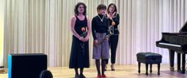 Na zdjęciu dwie kobiety. Jedna z nich brunetka z lokami ubrana w czarną suknię, a druga brunetka ubrana w szare spodnie i czarny golf. Obie trzymają po różyczce. Za nimi kobieta brunetka w czarnych spodniach i czarnej marynarce. Obok nich widać czarny fortepian.Tło obrazka w odcieniach beżu.
