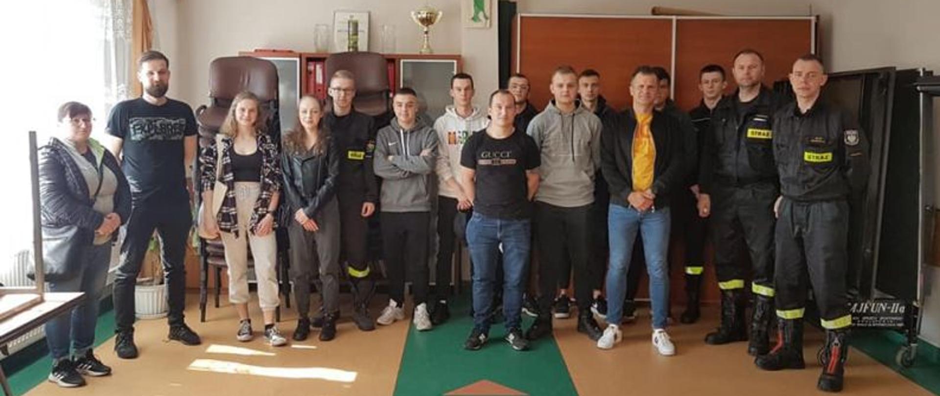 Zdjęcie kursantów, uczestników szkolenia. Zdjęcie grupowe. Widać na nim przyszłych druhów i druhny OSP. Po prawej stronie zdjęcia komisja egzaminacyjna składająca się ze strażaków w czarnych ubraniach koszarowych.