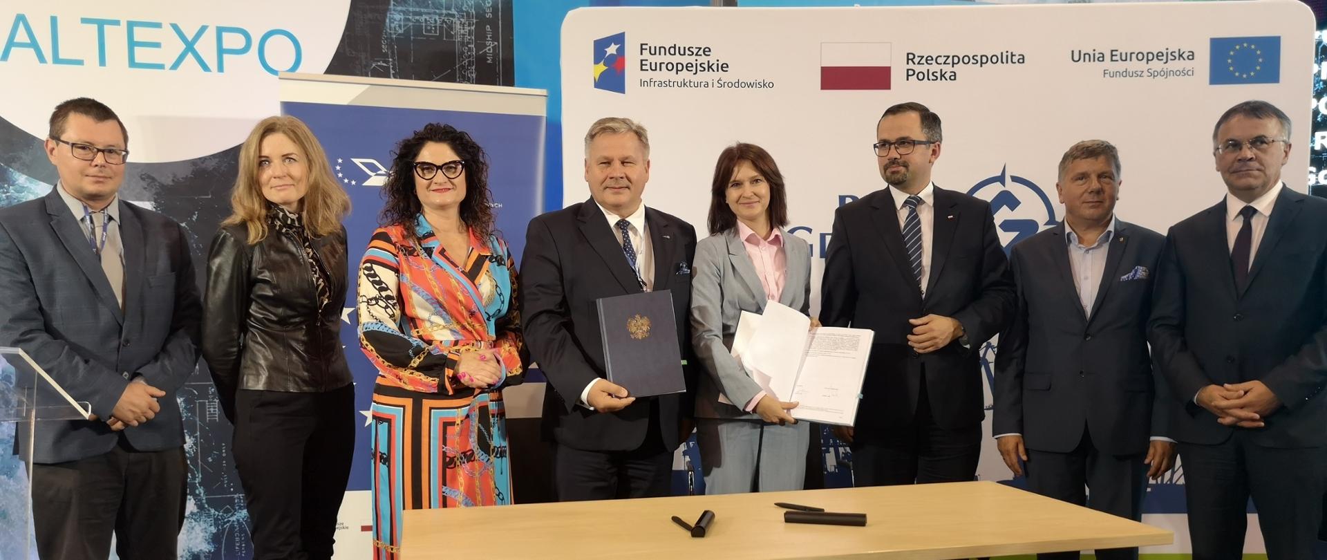 Za stołem stoi osiem osób, dwie z nich w ręku trzymają podpisane umowy. 