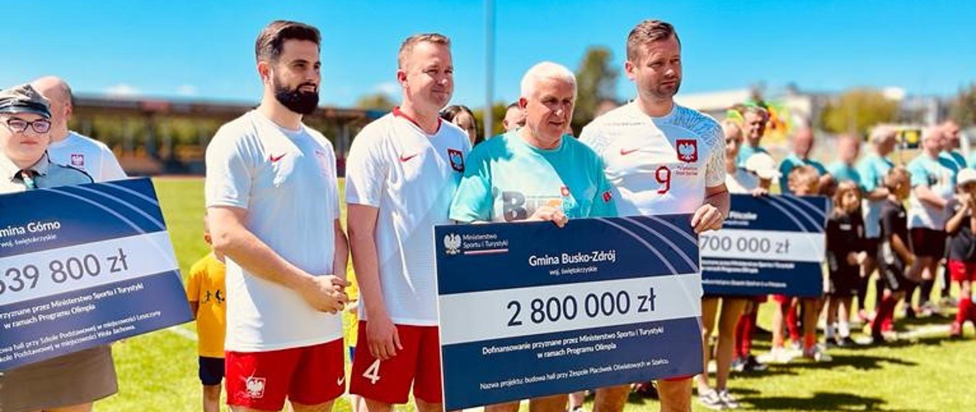 Minister Kamil Bortniczuk w stroju piłkarskim reprezentacji Polski stoi na murawie boiska. Wraz z drugim mężczyzną, którym jest burmistrz Waldemar Sikora z Buska-Zdroju, trzyma symboliczny czek dla Gminy Busko-Zdrój 