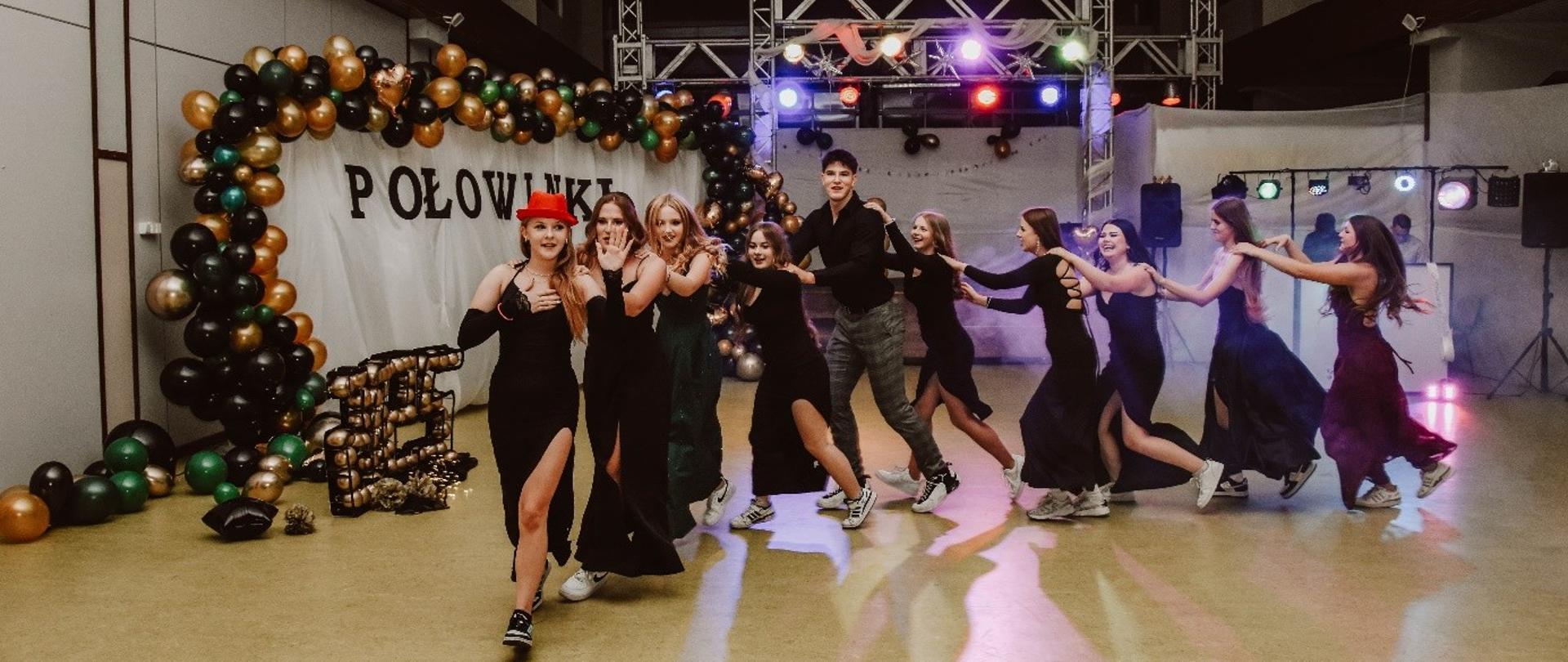 Grupa młodych ludzi, ubranych w eleganckie stroje wieczorowe, tańczy i bawi się podczas imprezy Połowinki na hali sportowej udekorowanej balonami w kolorach czarnym, złotym i zielonym.