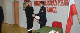 Świetlica komendy powiatowej. Na tle ściany, na której widnieje czerwony napis Komenda Powiatowa Państwowej Straży Pożarnej w Rawiczu i logo PSP, komendant powiatowy ściskając dłoń składa gratulacje jednemu z nowych strażaków. Z prawej strony toi stolik i stojak z flagami.