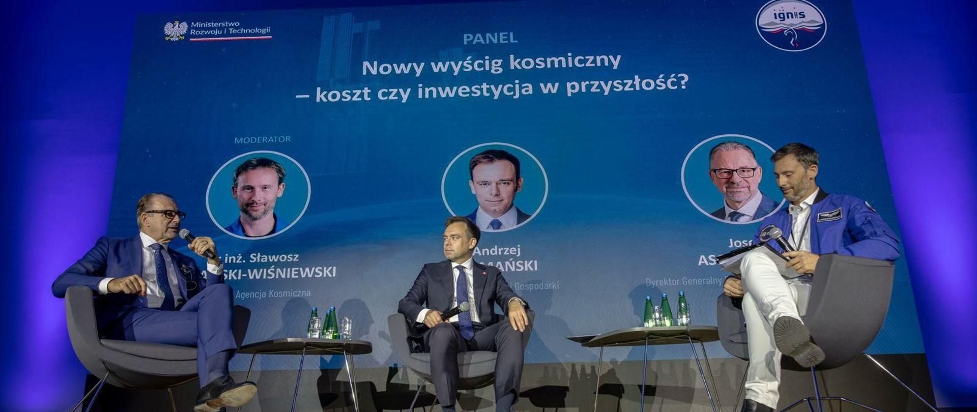 Zdjęcie z panelu na konferencji. Na podwyższeniu siedzą od lewej J. Asbacher, A. Domański, S. Uznański-Wiśniewski. W tle grafika z logotypami MRiT i IGNIS, zdjęciami mówców i tytułem panelu: Nowy wyścig kosmiczny - koszt czy inwestycja w przyszłość
